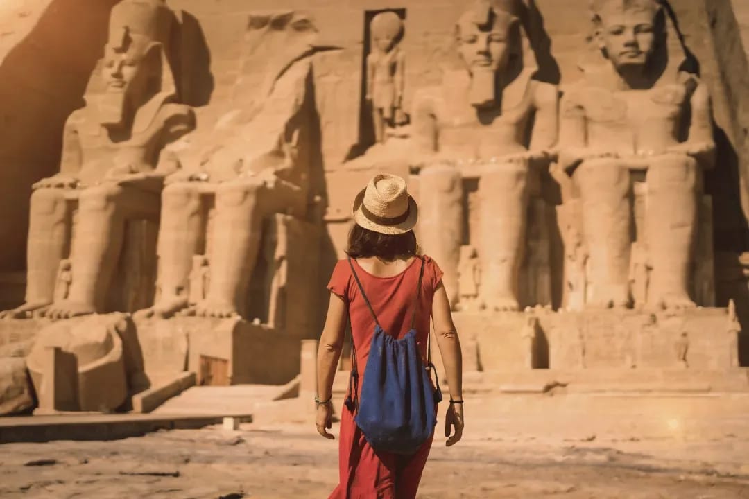 Woman in a sun hat and red dress facing the colossal statues of Abu Simbel