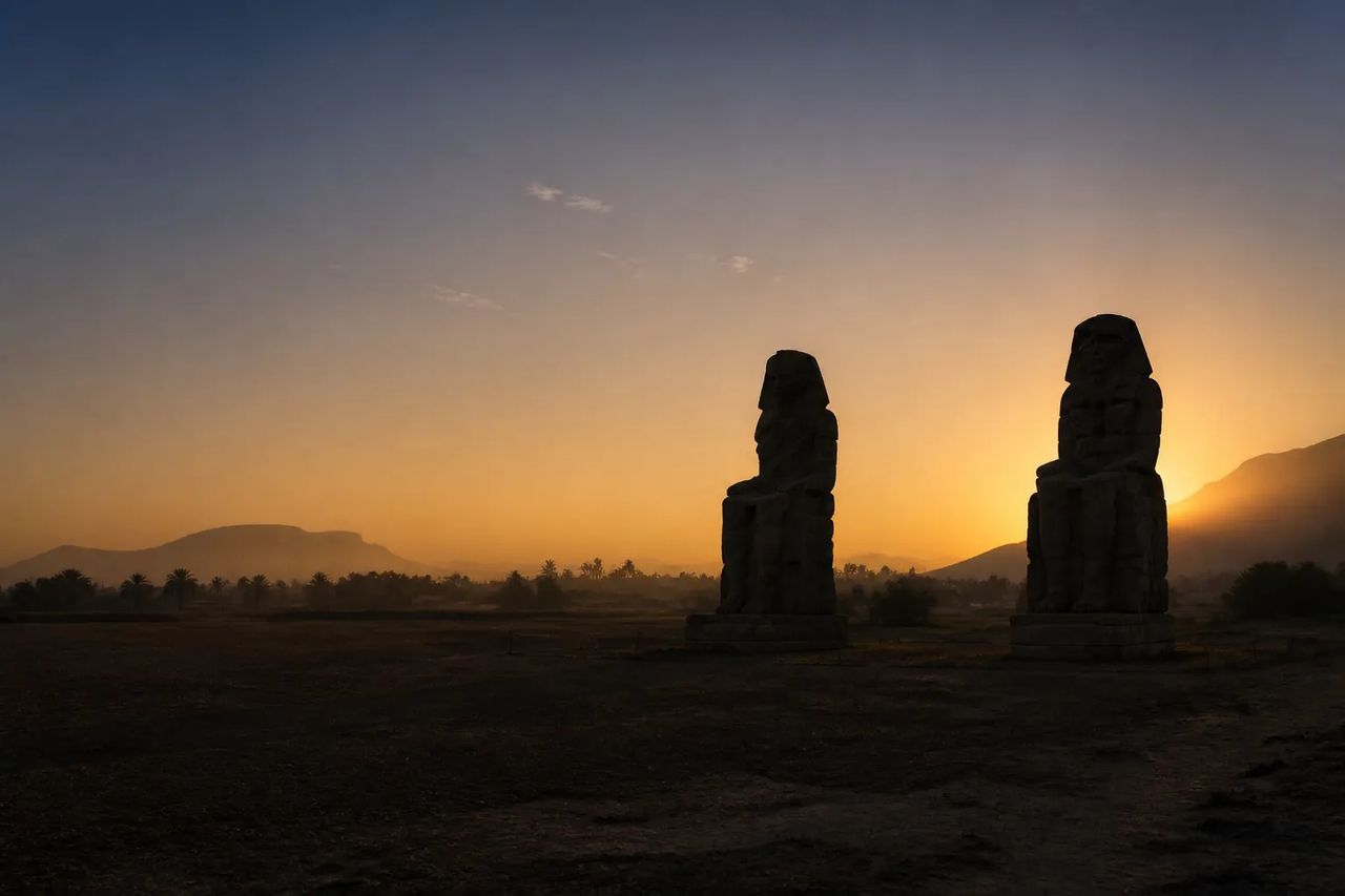 The Colossi of Memnon at sunrise, two seated stone figures silhouetted against a golden sky