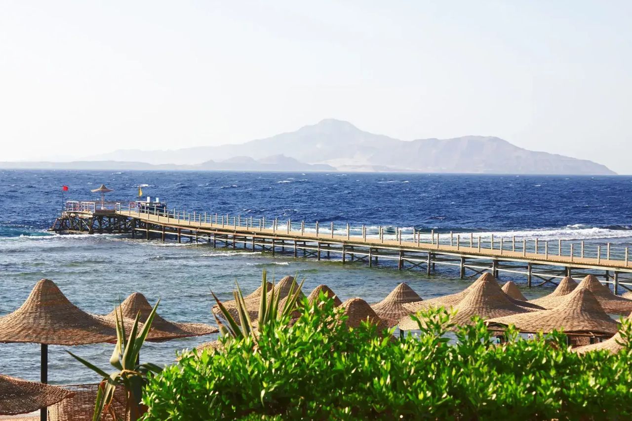 Wooden pier reaching into deep blue sea past beach parasols, with a mountainous island on the horizon
