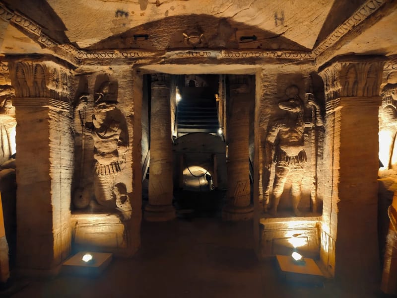 Lit rock-cut tomb chamber with carved figures and columns in the Kom el Shoqafa catacombs