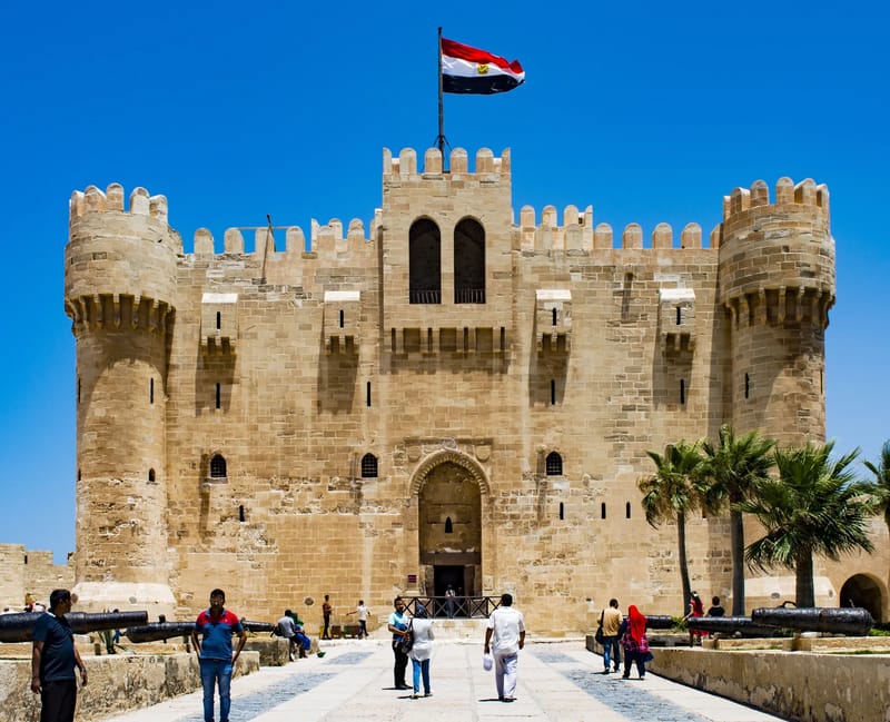 Main gate and towers of Qaitbay Citadel with a flag above and visitors on the approach