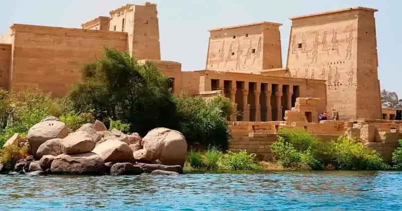 Temple pylons at Philae rising behind rocks and trees at the water edge