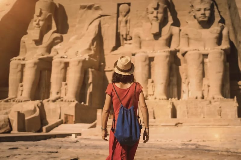 Woman in a sun hat and red dress facing the colossal statues of Abu Simbel